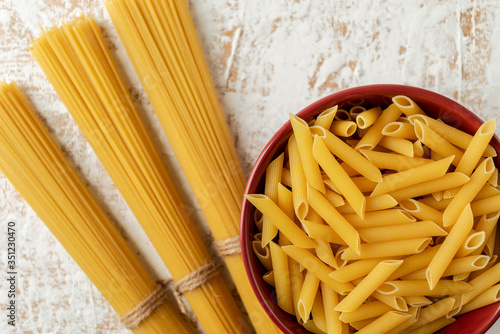 dry Penne pasta in a red ceramic bowl and bunches of spaghetti on a textured white-coffee background