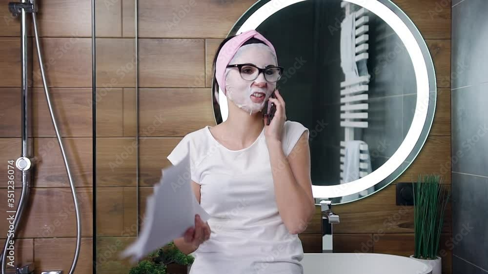 Front view of attractive satisfied young woman with white cosmetics mask on face which talking on phone and watching on financial reports Front view of attractive satisfied young woman with white cosmetics mask on face which talking on phone and watching on financial reports