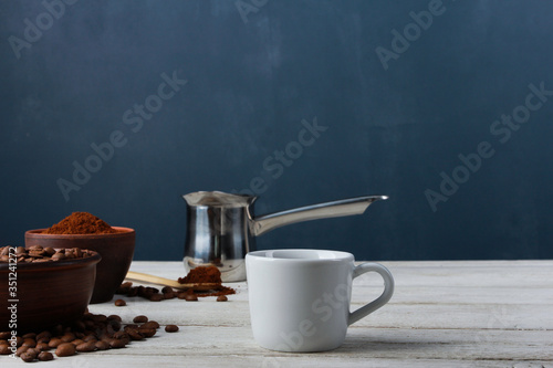 White coffee cup, Arabica beans in clay bowls, ground powder, metal Turkish pot on while table against dark blue wall. Side view, copy space. Coffee shop, morning, baristas workplace concept