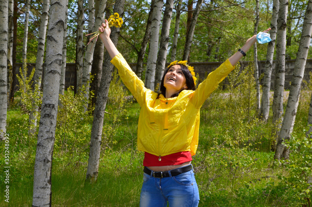 Happy cheerful woman in yellow jacket with wreath of dandelions,medical mask in hand and  bouquet of 
dandelions.she opened her arms to the sky. coronavirus pandemic end concept.