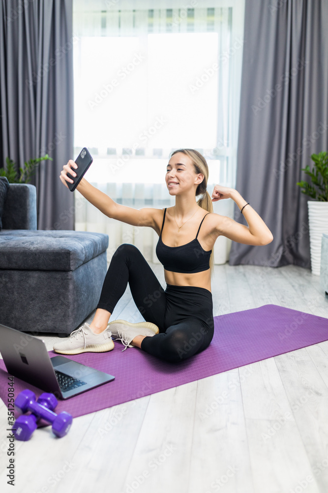 Obraz premium Young attractive sporty woman sitting on yoga mat making selfie with her mobile phone at home