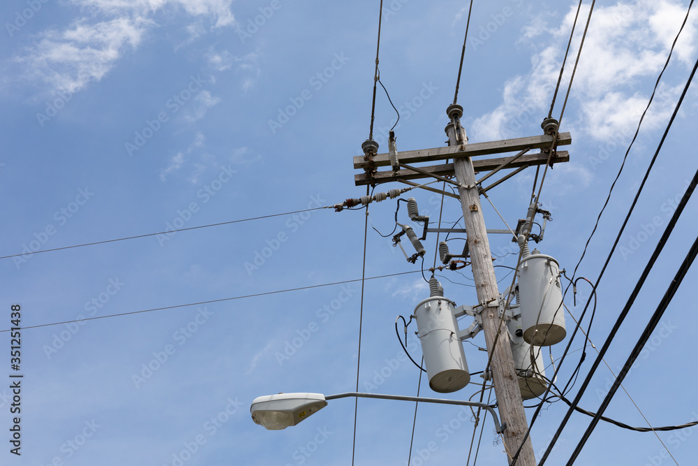 Top of a power pole with multiple transformers, insulators, wires, and a street light against a blue sky with clouds, creative copy space, horizontal aspect Top of a power pole with multiple transformers, insulators, wires, and a street light against a blue sky with clouds, creative copy space, horizontal aspect