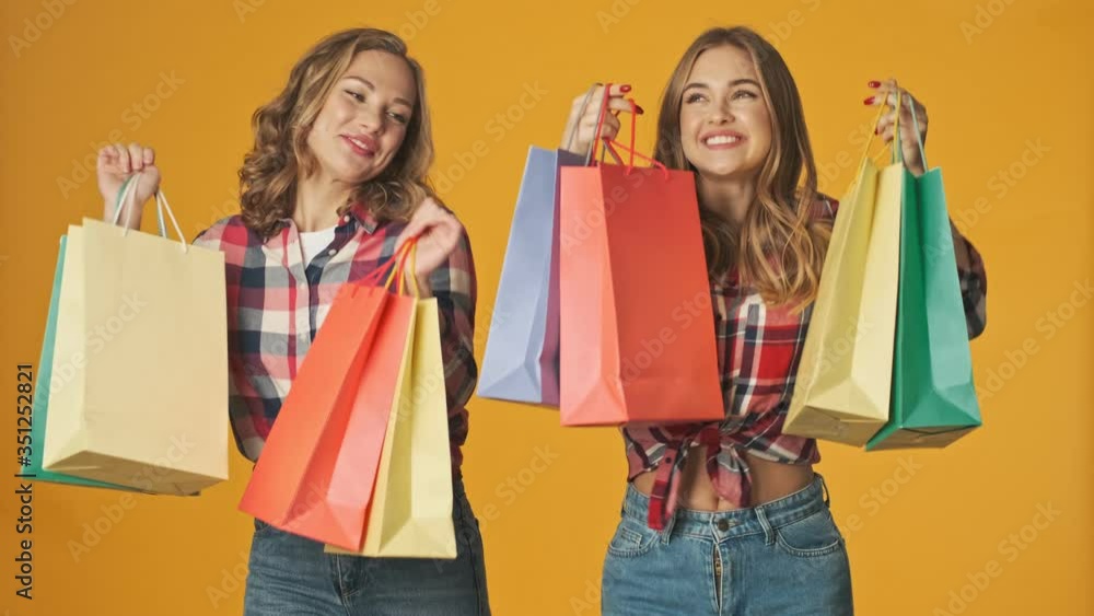 Young happy positive girls friends sisters isolated over yellow wall background holding shopping bags. Young happy positive girls friends sisters isolated over yellow wall background holding shopping bags.