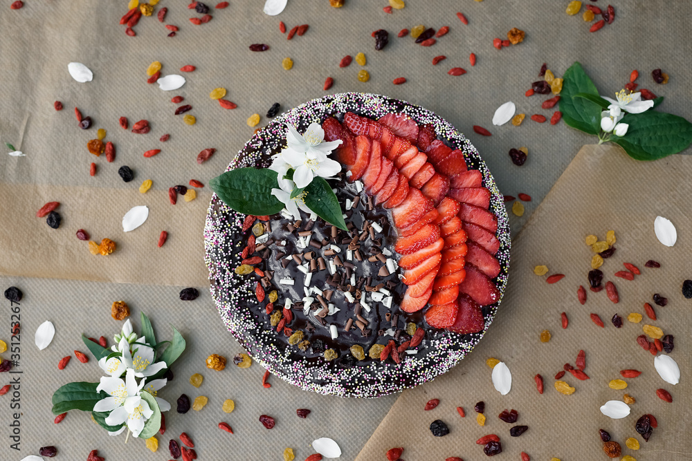Fototapeta premium Homemade strawberry cheese cake with raisins and dry fruits, triple chocolate curls, multi coloured sugar with chocolate topping on brown baking paper with dry fruits and flower of jasmine. Flat lay.
