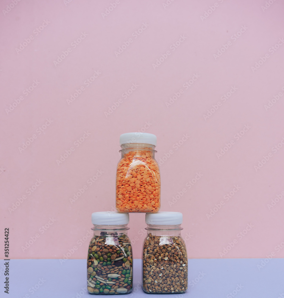 Small bottles of dry cereals on pink and blue background. Different types of cereals and legumes, close up. Set of whole grains - buckwheat, red lentil, beans, peas, red beans. Copy space.