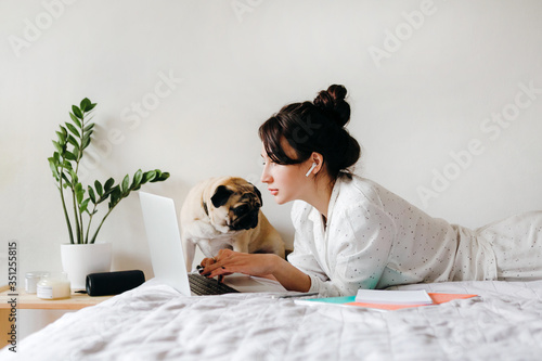 Young beatiful girl is working with laptop on bed near a dog at home in isolation. Home office. Concept of working and studying in isolation. photo