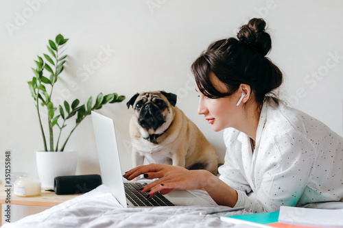 Young beatiful girl is working with laptop on bed near a dog at home in isolation. Home office. Concept of working and studying in isolation.