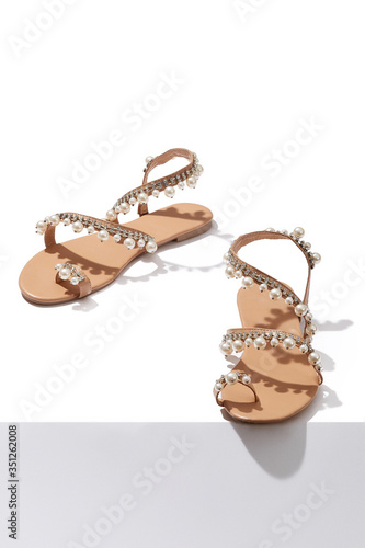 Detailed shot of beige flat sandals with toe ring and decorated with rhinestones and pearls. The pair of summer footwear is isolated on the white surface. 