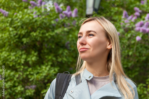 Close-up face a blond girl background green bushes of lilac with pensive confident eyes looking up and side with blue eyes. Calm and confidence in the expectation of a new life during summer holidays.