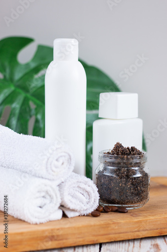Handmade coffee scrub in a glass jar on a light wooden table. White towels rolled up and bottles with cosmetic. The concept of home care and spa treatments.