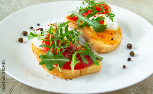 delicious poached eggs on crispy slices of wheat bread with slices of tomato, arugula, spices and black pepper on a white plate on a concrete background