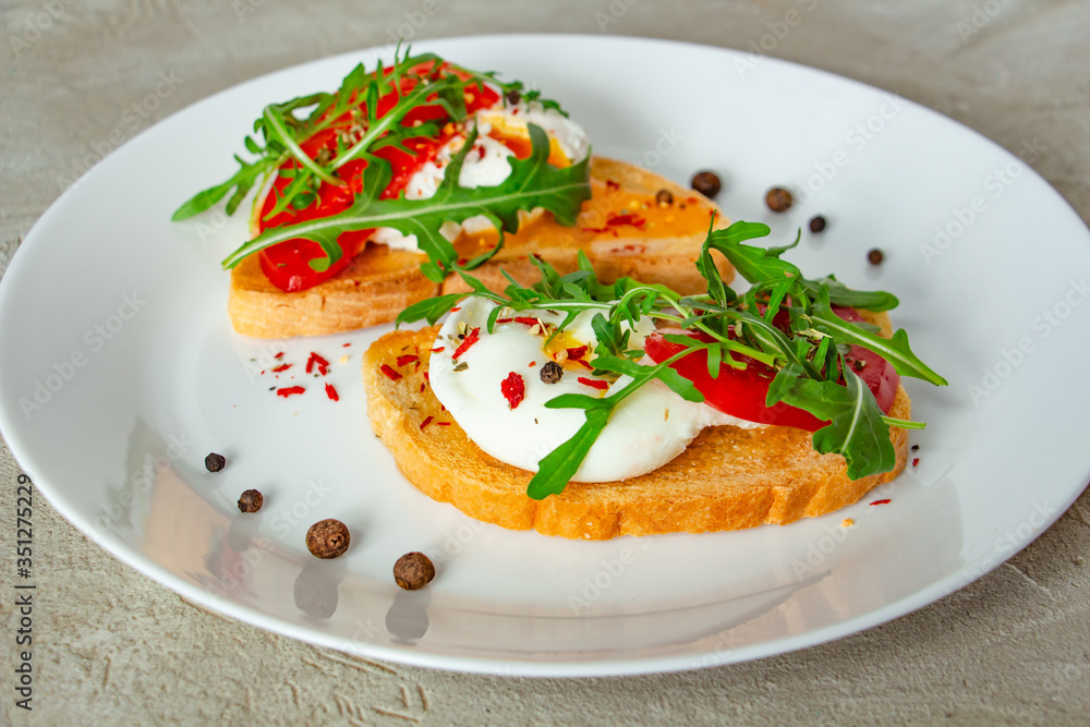 delicious poached eggs on crispy slices of wheat bread with slices of tomato, arugula, spices and black pepper on a white plate on a concrete background