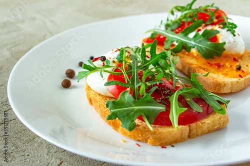 delicious poached eggs on crispy slices of wheat bread with slices of tomato, arugula, spices and black pepper on a white plate on a concrete background