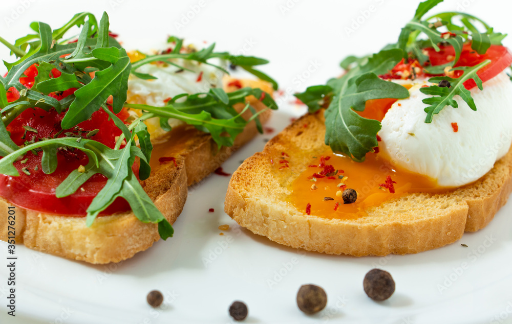 delicious poached eggs on crispy slices of wheat bread with slices of tomato, arugula, spices and black pepper on a white plate on a white background
