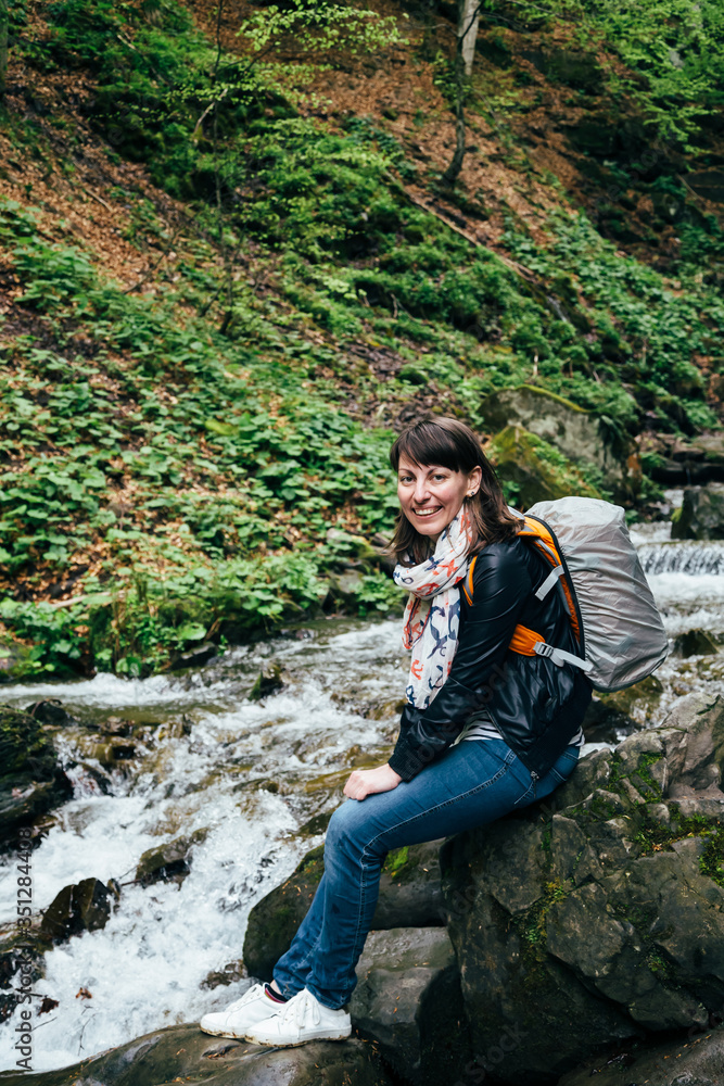 Naklejka premium Happy smiling tourist girl in blue jeans, striped shirt and black jacket with backpack near to the cold fresh river at the waterfall Shypit, cascade in Pylypets in the forest. Carpathian Mountains