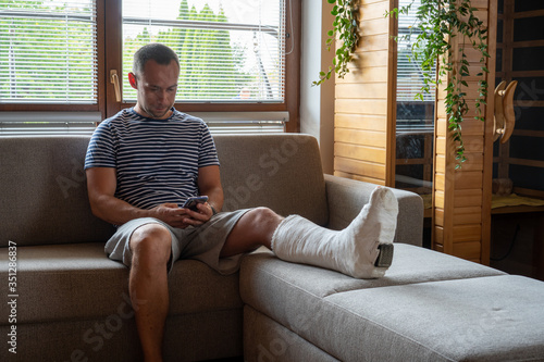 Man with broken leg in cast using mobile phone while sitting in sofa at home