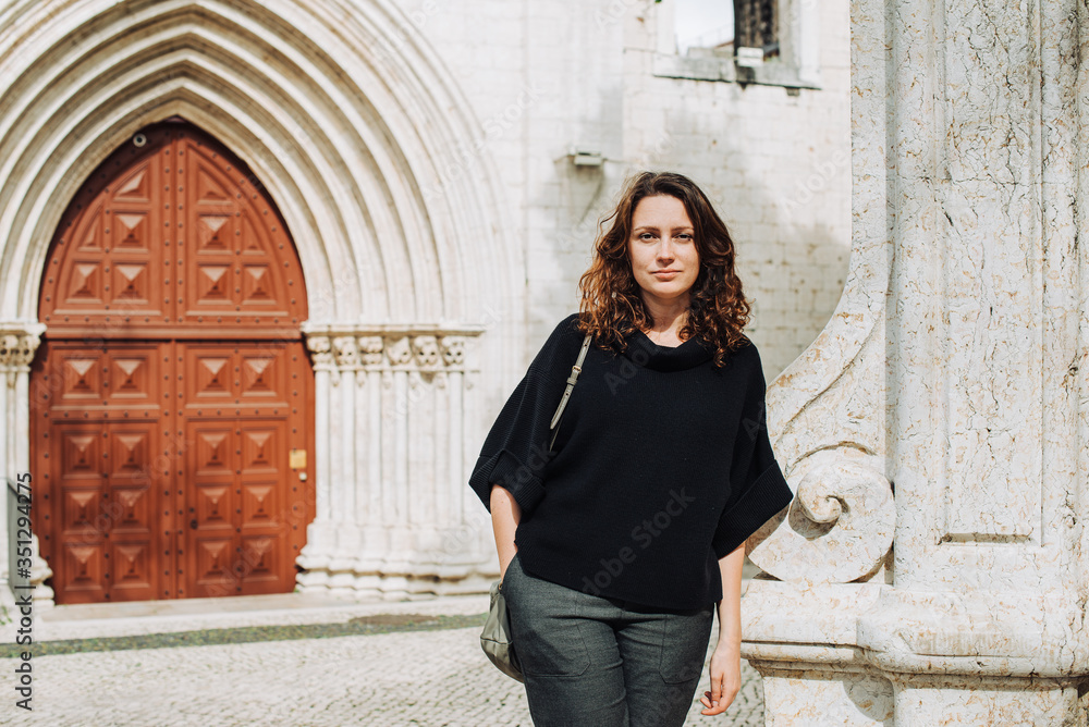Young Caucasian woman standing at portal of a gothic church wearing dark casual clothing and looking at camera. Lisbon as a travel destination concept. Architecture elements, entrance group context 
