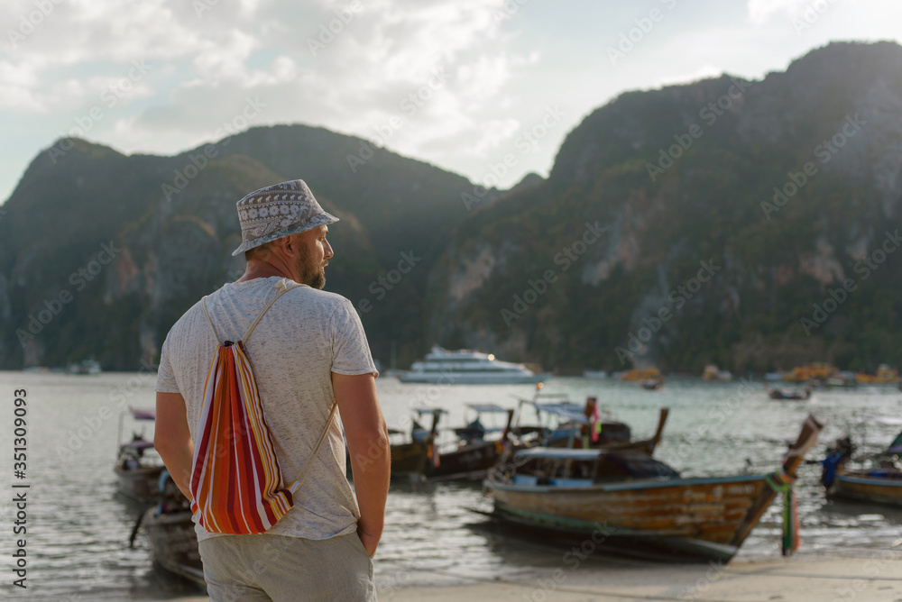 Young male tourist in a hat and with a backpack on a Thai beach on a background of traditional Thai boats.