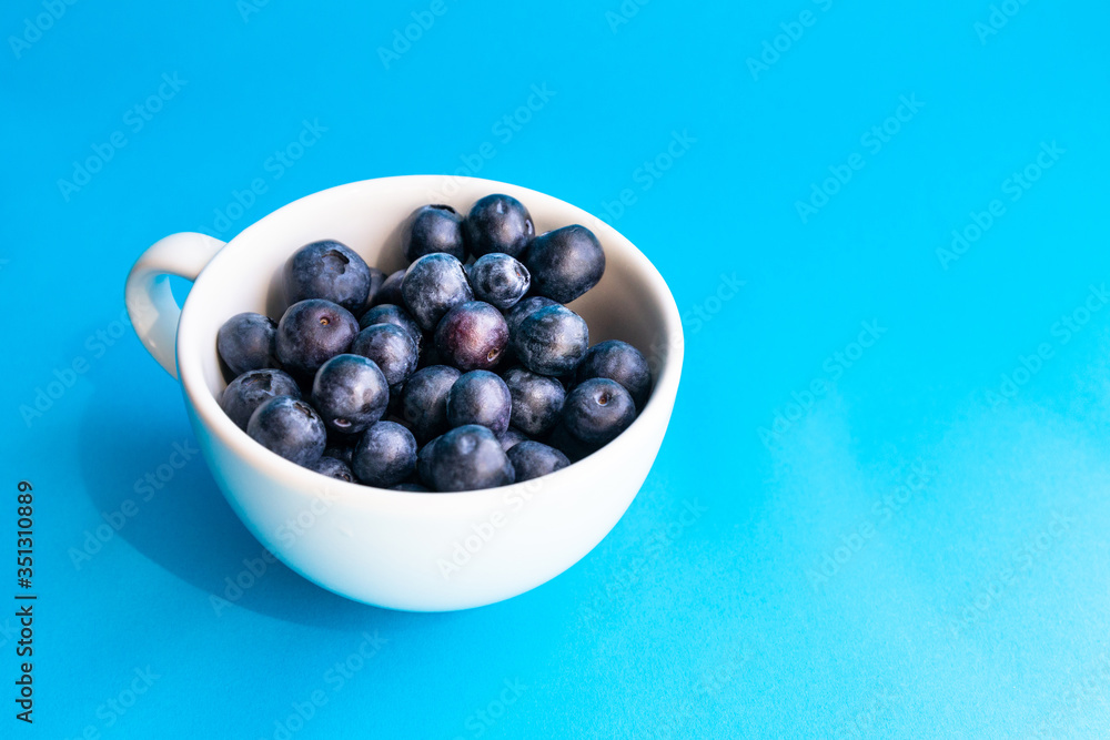 Ripe blueberry and green leaves in white cup on blue horizontal background Ripe blueberry and green leaves in white cup on blue horizontal background