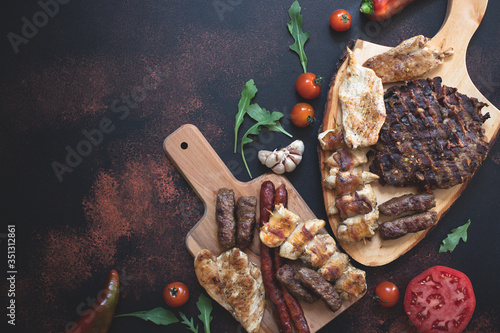 Various grilled meats served on wooden cutting boards with fresh vegetables and bread on a dark rustic background. Traditional Serbian and Balkan bbq meat called rostilj. Flat lay. Copy space photo