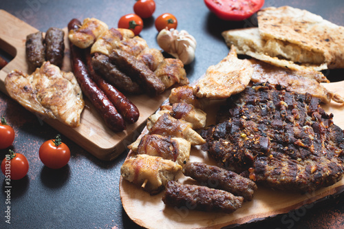 Various grilled meats served on wooden cutting boards with fresh vegetables and bread on a dark rustic background. Traditional Serbian and Balkan bbq meat called rostilj. Close-up. Copy space  photo