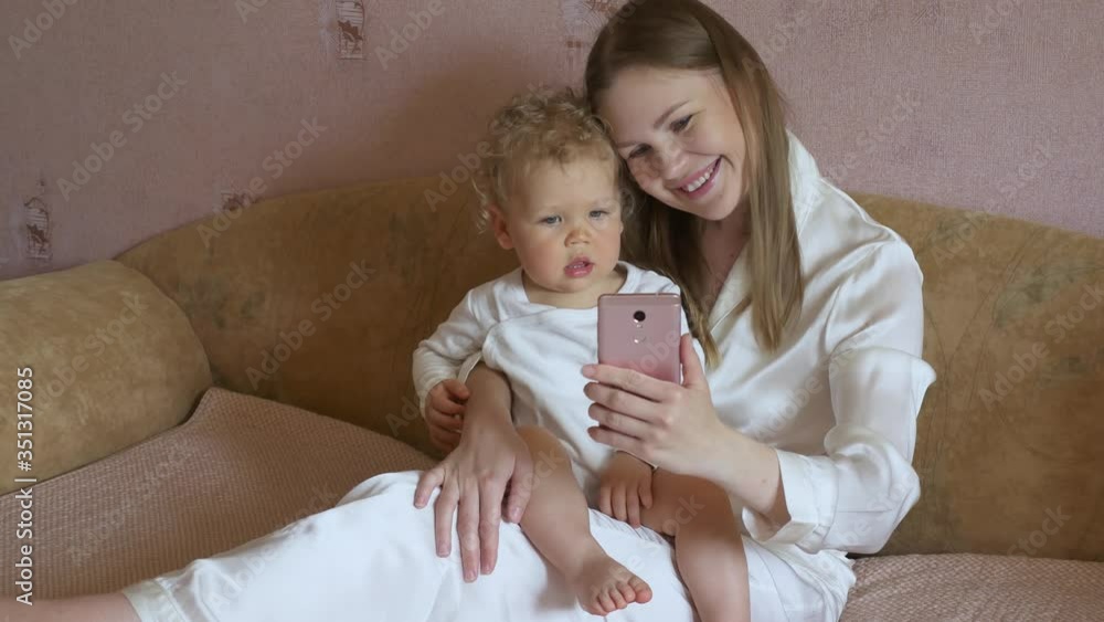 Beautiful attractive mother with a cute baby sits at home on the couch and gazes cheerfully at a mobile phone. Beautiful attractive mother with a cute baby sits at home on the couch and gazes cheerfully at a mobile phone.