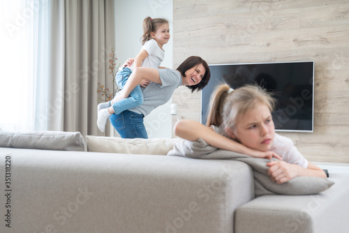 Preteenage sad girl sitting on a couch while mother having fun with sister, jealousy concept photo