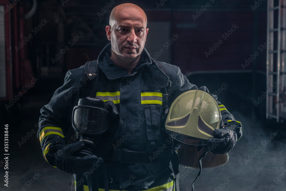 Fototapeta premium Fireman standing confident holding helmet and wearing firefighter turnouts. Portrait of a fireman with dark background with smoke and blue light.