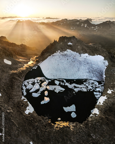 Aerial view of small glacier and adjacent lagoon lake with floating ice shelfs during sunrise, with mountains peaking over the fog in the background. Ticino, Switzerland photo
