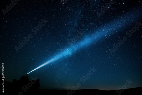 Night landscape of a man with a powerful military flashlight illuminating the starry sky leaving a beam of light