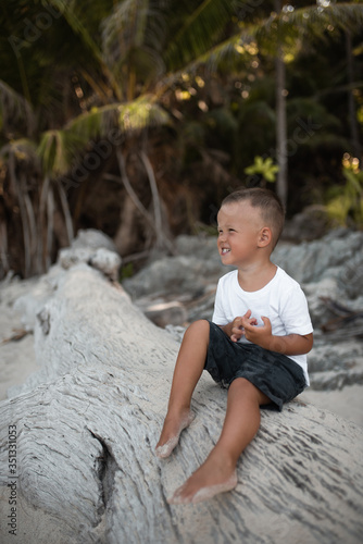 Fototapeta Naklejka Na Ścianę i Meble -  Happy smile european blond baby boy have rest and sit on white sand beach. . white cotton clothes. t-short.Thailand. palms. rocks