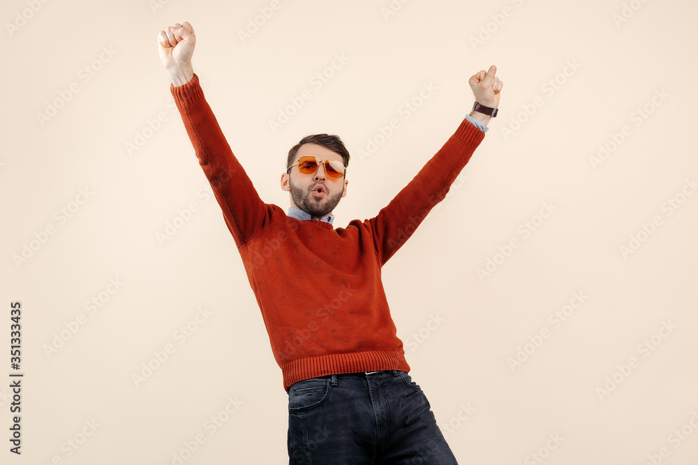 Cheerful bearded man in casual outfit wearing sunglasses feelling happy on beige background. Success and winner.