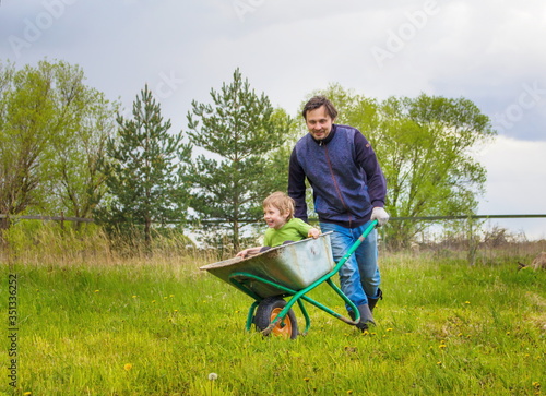 Man rolls his son in a garden cart in the garden. The son laughs and rejoices.