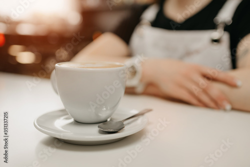 A female barista waits with a cup of fresh coffee with a saucer and spoon for a client in a cafe. A barista with a cup of latte in a coffee shop.