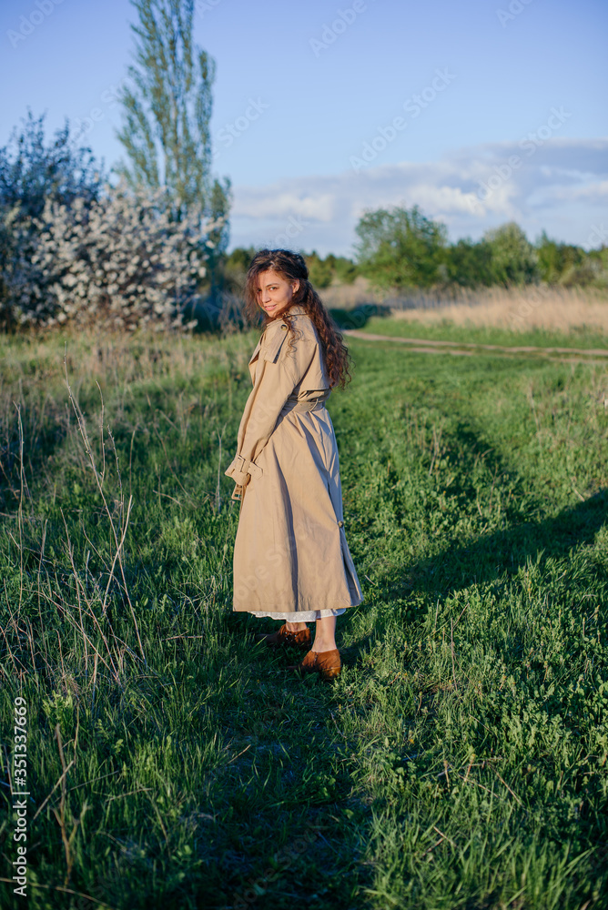 A girl with long hair and a beige trench coat walks along a country road, woman walking in the countryside