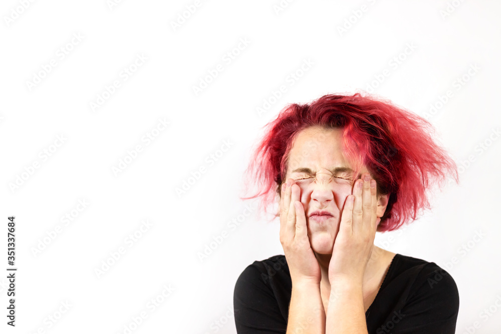 A close-up of a sad girl with disheveled red dyed hair squeezed her eyes shut and pressed her hands to her face. On white background. An emotion of frustration. Bad hair day.