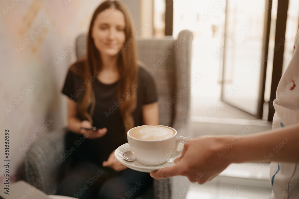 A close-up photo of a latte which a female barista holds out to a girl in a coffee shop. A woman with long hair working remotely on a laptop keeps social distance grabs a cup of coffee in a cafe. A close-up photo of a latte which a female barista holds out to a girl in a coffee shop. A woman with long hair working remotely on a laptop keeps social distance grabs a cup of coffee in a cafe.