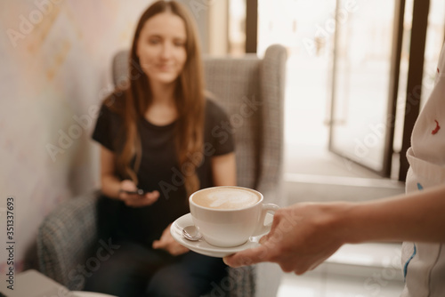A close-up photo of a latte which a female barista holds out to a girl in a coffee shop. A woman with long hair working remotely on a laptop keeps social distance grabs a cup of coffee in a cafe.