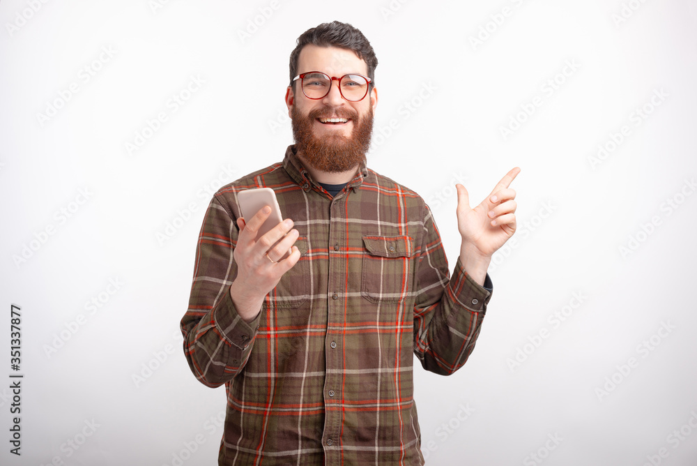 Handsome bearded man wearing glasses is holding a phone and pointing aside on white. Handsome bearded man wearing glasses is holding a phone and pointing aside on white.