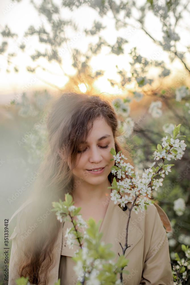 Fototapeta premium A young girl with long hair in a beige trench coat and white dress next to a flowering tree, girl's face in flowers