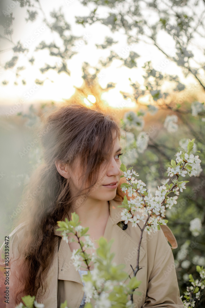 A young girl with long hair in a beige trench coat and white dress next to a flowering tree, girl's face in flowers A young girl with long hair in a beige trench coat and white dress next to a flowering tree, girl's face in flowers