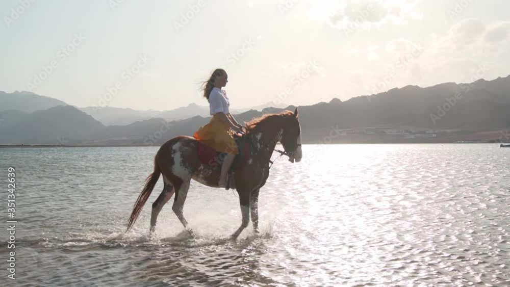 Caucasian blonde girl riding a horse through the water at sunrise slow motion Caucasian blonde girl riding a horse through the water at sunrise slow motion