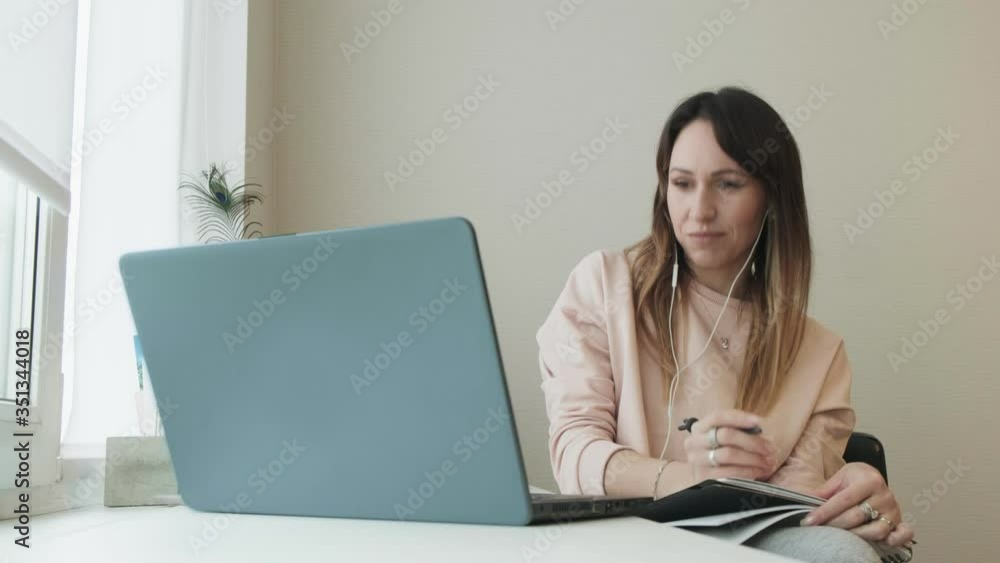 Thoughtful smiling caucasian woman wearing headset sitting at the table on video call with work colleagues working on laptop computer takes notes in notebook Self-isolation at Pandemic Thoughtful smiling caucasian woman wearing headset sitting at the table on video call with work colleagues working on laptop computer takes notes in notebook Self-isolation at Pandemic