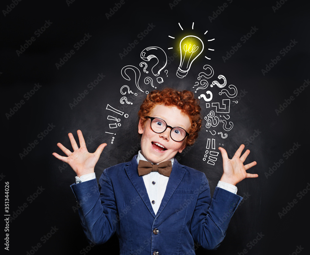 Redhead boy is is mischievous against blackboard background with lightbulb and question marks. Brainstorming and idea concept Redhead boy is is mischievous against blackboard background with lightbulb and question marks. Brainstorming and idea concept