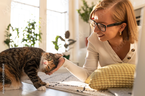 Smiling woman freelancer in glasses lies on the carpet in living room at home, stroking and plays with cat. Cat interferes with work on laptop. Love pets. photo