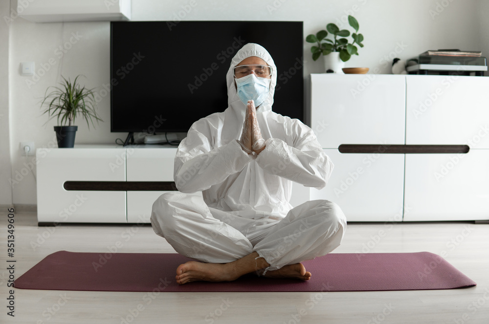 Young man in medical mask and in protective suit is practicing yoga on a mat at home. Stay home during COVID-19 quarantine concept. Yoga at home online. Coronavirus pandemic. Healthy lifestyle. Young man in medical mask and in protective suit is practicing yoga on a mat at home. Stay home during COVID-19 quarantine concept. Yoga at home online. Coronavirus pandemic. Healthy lifestyle.