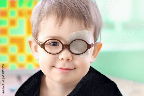 Obraz na plátně A little boy wearing glasses and an eye patch (plaster, occluder) undergoes a hardware vision treatment to prevent amblyopia and strabismus (squint, lazy eye)