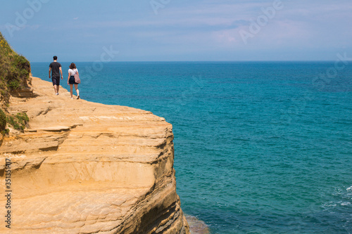 Couple traveling together on sand cliff edge on blue sea and sky background Sidari, Greece. Young family of travelers on hill with stunning views of the ocean. Summer lifestyle concept.