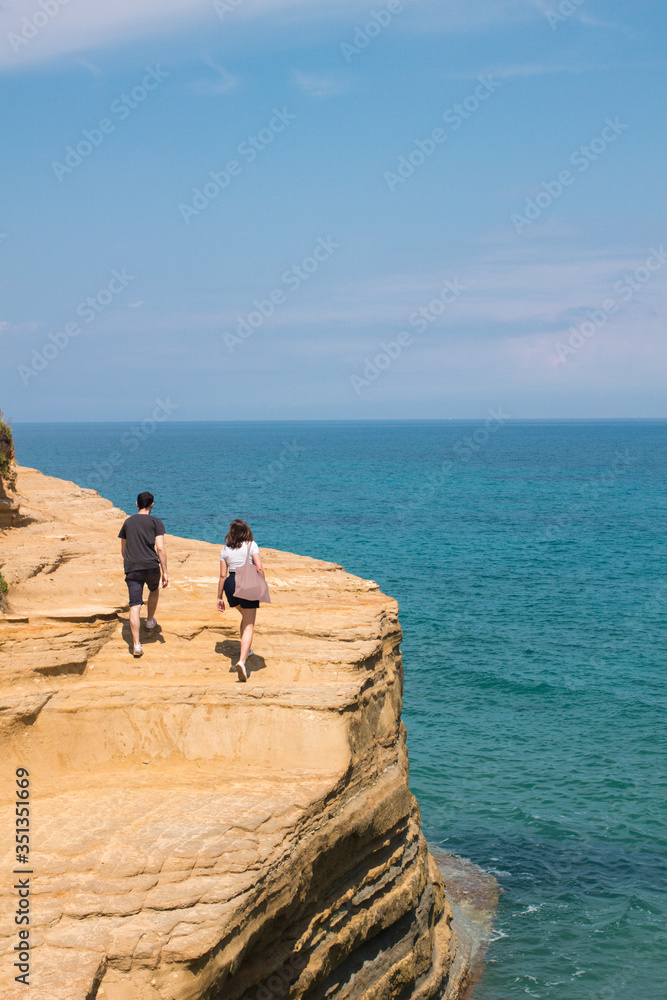 Millennial couple in summer outfit rising walking on sandy cliff rock edge on blue emerald crystal clear sea ocean background in sunny day.View from back. Summer travel vacation concept. 