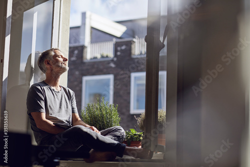 Mature man sitting at the window at home with closed eyes photo